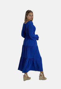 Kongeblå, langærmet maxikjole med lagdelt design og smocktalje. Bruges med beige kilehælssandaler. Glat stoftekstur.