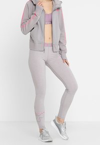 Donna che indossa leggins atletici grigio chiaro, giacca a zip abbinata, reggiseno sportivo e sneakers argento, in piedi con una gamba leggermente in avanti.