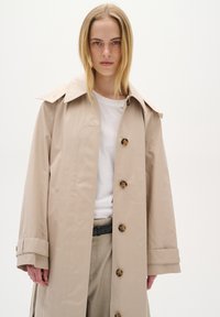 Beige trenčkot s širokým límcem, velkými knoflíky a mírně oversized střihem. Materiál vypadá hladce a je voděodolný.