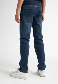 Jeans en denim bleu sombre avec une coupe droite, poches arrière visibles et texture délavée. Associés à des baskets blanches.