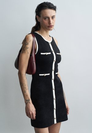 Rochie neagră fără mâneci, dintr-un material texturat, cu nasturi albi în față și accente de buzunare. Modelul ține o geantă burgundy, stând pe un fundal deschis.