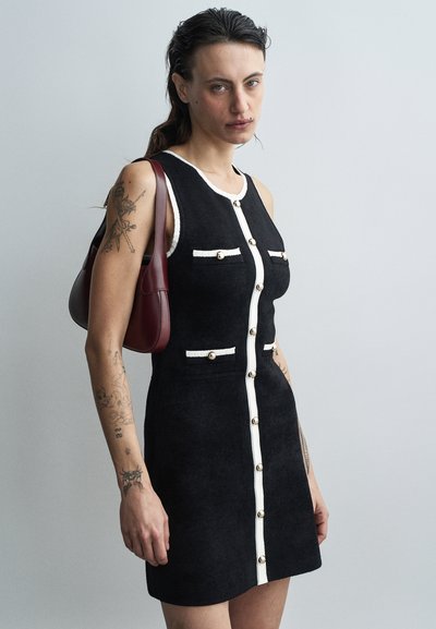 Robe sans manches noire avec un tissu texturé, boutons blancs sur le devant et poches en accent. Le modèle tient un sac bordeaux, se tenant devant un fond clair.
