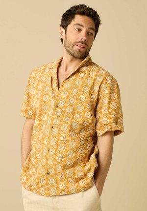Man draagt geel overhemd met korte mouwen en witte bloemenprint, handen in de zakken, kijkt zijwaarts tegen een beige achtergrond.