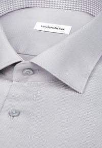 Chemise habillée gris clair texturée avec un col pointu, deux boutons visibles, et une étiquette blanche portant l'inscription "seidensticker" à l'intérieur du col.
