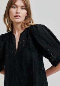 Blouse noire texturée avec des manches bouffantes, un décolleté en V avec des détails découpés, et un design en tissu doux et superposé.
