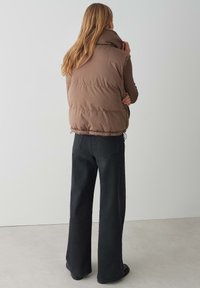 Braune, gepolsterte Weste mit hohem Kragen, die eine glatte Textur und einen elastischen Saum aufweist, kombiniert mit dunklen, weiten Hosen in einer legeren Umgebung.