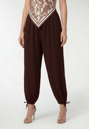 Donna che indossa pantaloni marrone scuro larghi con lacci alla caviglia abbinati a sandali dorati con tacco e una maglietta fantasia.