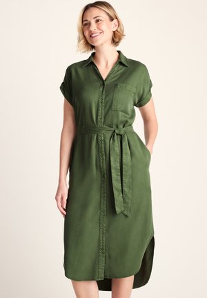 Woman smiling wearing a short-sleeve olive green button-down dress with a collar, chest pocket, tied waist belt, and side slit.