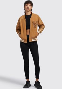 Pruun bomber-jakk, millel on tiigri logo, kontrastsed valged triibud varrukatel ja alläärdel ning sile, läikiv tekstuur. Must topp ja legginsid.