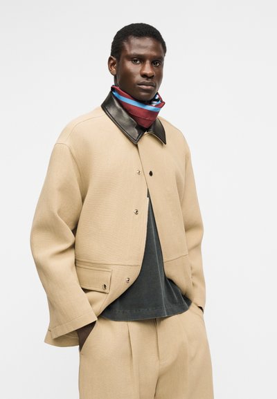 Άνδρας με μπεζ σακάκι και παντελόνι, μαύρο πουκάμισο και κόκκινο-μπλε ριγέ κασκόλ, στέκεται με τα χέρια στις τσέπες μπροστά σε μονόχρωμο φόντο.