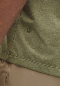 Oliwkowa koszula z krótkim rękawem, wykonana z miękkiego materiału, z delikatnym logo i prostym dołem. Ujęta na beżowym tle.