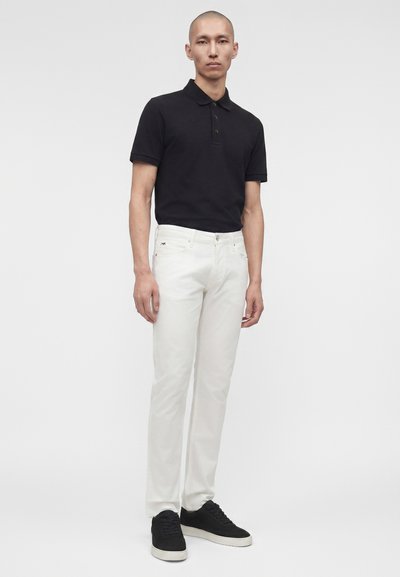 Άνδρας με μαύρο πόλο, λευκά τζιν και μαύρα αθλητικά παπούτσια, στέκεται μπροστά σε απλό λευκό φόντο.