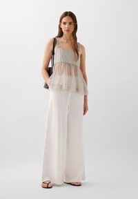 Top trasparente grigio con strati e spalline a volant abbinato a pantaloni bianchi larghi. La modella porta una borsa nera; design lungo fino al pavimento, vestibilità comoda.