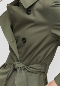 Trenchcoat in Olivegrün aus glattem Stoff, mit doppelreihiger Front, schwarzen Knöpfen und einem gebundenen Gürtel in der Taille.