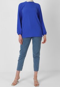 Maglia blu a maniche lunghe con scollo rotondo e polsini elastici, abbinata a jeans dritti azzurri. Calzature sono sandali con tacco aperto.