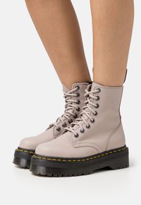 Botas de combate de cuero rosa claro con punta redonda, suelas de goma negra gruesa con un pespunte amarillo y detalles de cordones negros.