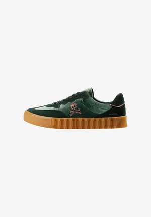 Sneaker verde in suede con suola in gomma, caratterizzata da un design laterale di teschio e tibie e cuciture tonali. Lacci neri piatti e punta arrotondata.