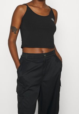 Kobieta w czarnym topie odsłaniającym brzuch i czarnych spodniach typu cargo, z rękami w kieszeniach, pokazująca tatuaż pantery na górnej części lewej ręki.