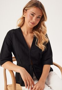 Svart wrap-top med djup V-ringning, korta ärmar och knytning vid midjan. Tyget verkar lätt och texturerat.