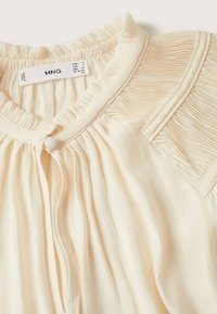 Blusa color beige chiaro con texture plissettata sulle spalle, scollatura rotonda, chiusura con bottoni e tessuto morbido. Etichetta visibile all'interno del colletto.
