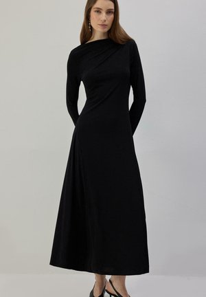 Kvinde iført en lang sort asymmetrisk kjole med den ene skulder dækket, stående foran en ensfarvet lys grå baggrund.