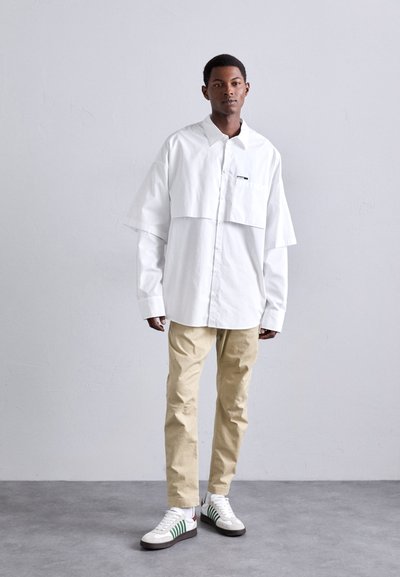 Weißes, übergroßes Hemd mit Knopfleiste und cropped Design, kombiniert mit beigen Hosen und weißen Sneakers mit grünen Streifen. Minimalistischer Stil.