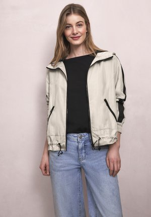 Junge Frau trägt eine hellbeige Reißverschlussjacke über einem schwarzen Shirt und hellblauen Jeans, steht vor einem einfarbigen hellen Hintergrund.