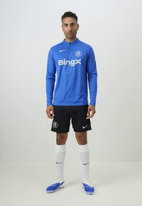Maglia da calcio a maniche lunghe blu con loghi bianchi, pantaloncini neri, scarpe da calcio blu e calzini bianchi. Il materiale sembra atletico e traspirante.
