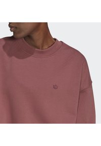 Person som bär en dammrosafärgad rundhalsad sweatshirt med en liten broderad logotyp på vänster bröst, visad från axlarna till hakan.