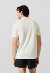 Camisa polo de manga corta de color beige claro con tejido texturizado y un logo negro en el cuello trasero. Combinada con pantalones cortos negros, vista desde atrás.