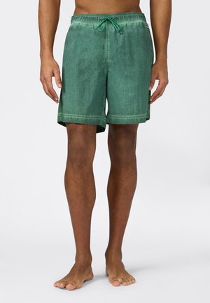 Mężczyzna model ubrany w zielone kąpielówki na sznurek, stojący boso na jednolitym jasnoszarym tle.