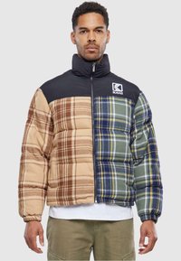 Puffervest med en blanding af ternede mønstre i brun, beige og grøn. Har en sort høj krave og lynlås lukning. KANI-logo foran.