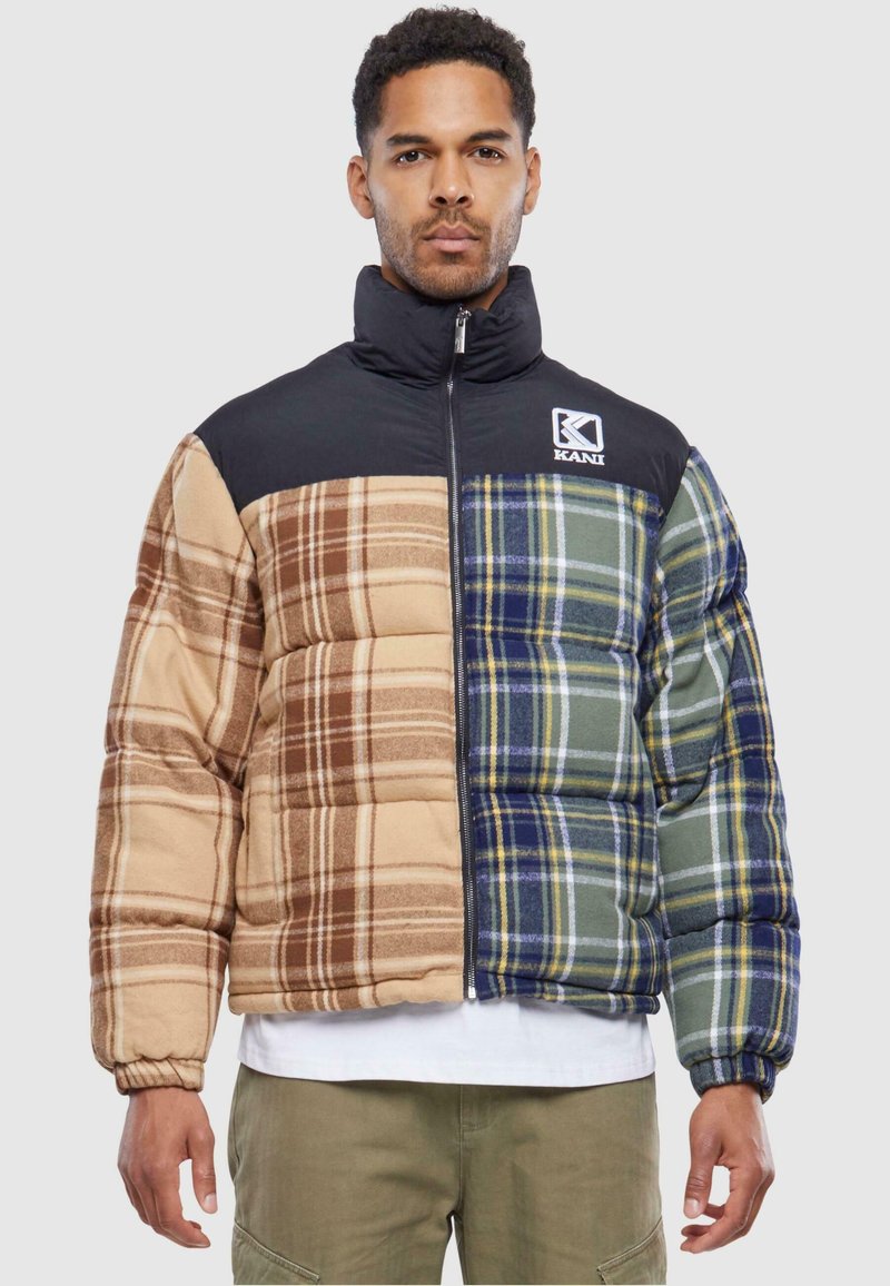 Puffervest med en blanding af ternede mønstre i brun, beige og grøn. Har en sort høj krave og lynlås lukning. KANI-logo foran.