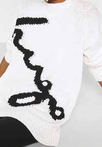 Maglione bianco lavorato a maglia con un design astratto nero. Il tessuto è texturizzato con orlo a coste e maniche corte.