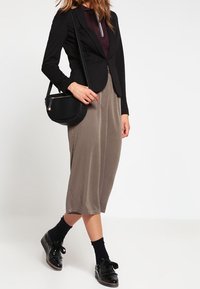 Blazer negro ajustado sobre una blusa color burdeos oscuro, acompañado de una falda midi de taupe suave. Los accesorios incluyen un pequeño bolso cruzado negro y zapatos de charol.
