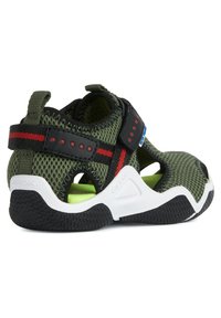 Sandal in rete verde con accenti neri e rossi, fascetta in Velcro, suola bianca e ritagli circolari per la ventilazione. Suola in gomma testurizzata.