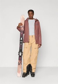 Žena u skijaškoj jakni i hlačama drži skiju okomito, obučena u skijaške čizme, stoji ispred jednostavne pozadine.