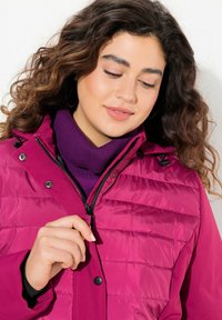 Magenta insulated jacket with horizontal stitching, zip closure, and adjustable hood. Worn over a purple turtleneck with ribbed texture.