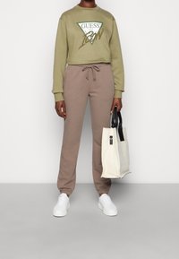 Sudadera verde olivo con el logo "GUESS", pantalones de chándal marrones, zapatillas blancas y una bolsa de lona beige con detalles negros.