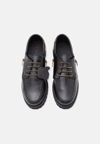 Chaussures en cuir noir avec bout rond, accents beige et lacets décoratifs. Présentent une étiquette logo attachée aux lacets. Texture lisse et semelle robuste.