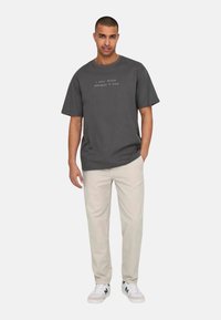 Gray t-shirt with short sleeves and a round neckline, featuring white text that reads "I ONLY ACCEPT APOLOGIES IN CASH." Paired with beige pants and sneakers.
