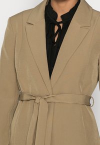 Béžový blejzr s texturovaným povrchem, s páskem v pěkném pasu, ostrými klopami a saténovým leskem. Nosený přes černou top s volánky.