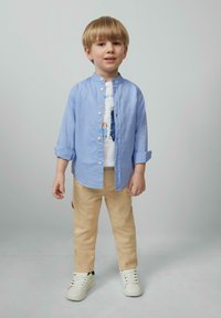 Lichtblauw overhemd met knopen over een witte graphic t-shirt, gecombineerd met beige broek en witte sneakers. Handen ontspannen langs de zij, lachend.