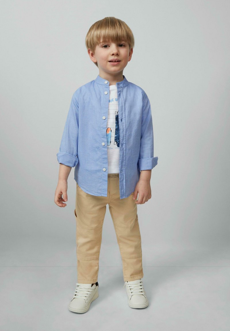 Lichtblauw overhemd met knopen over een witte graphic t-shirt, gecombineerd met beige broek en witte sneakers. Handen ontspannen langs de zij, lachend.