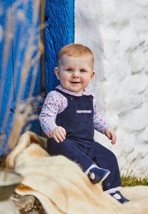 Kind in marineblauen Corduroy-Latzhosen mit einem blumigen Langarmhemd, sitzt auf einer beigen Decke vor einer strukturierten weißen Wand und einer blauen Tür.