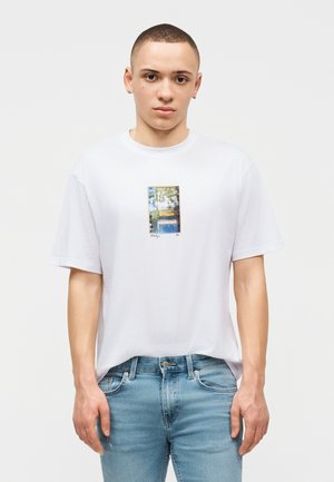 Νέος άνδρας με κοντά μαλλιά, που φοράει λευκό T-shirt με μικρό ορθογώνιο εκτυπωμένο φωτογραφικό σχέδιο και ανοιχτόχρωμο μπλε τζιν, στέκεται μπροστά σε απλό φόντο.