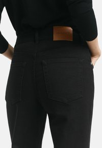 Jean en denim noir à taille haute, avec deux poches arrière et une étiquette en cuir beige portant la marque "GANT" au niveau de la taille.