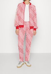 Röd och vit mönstrad träningsdräkt med en bomberjacka med dragkedja och matchande byxor. Har mjukt tyg, elastiska muddar och dragsko i midjan.