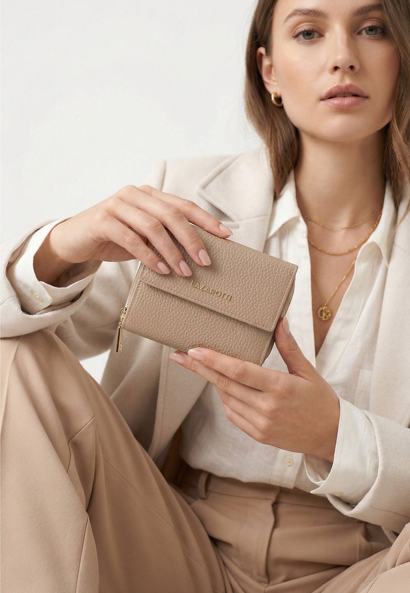 Vrouw in beige pak die een gestructureerde beige portemonnee vasthoudt met "LAZAROTTI" in reliëf op de klep, die gouden sieraden draagt.