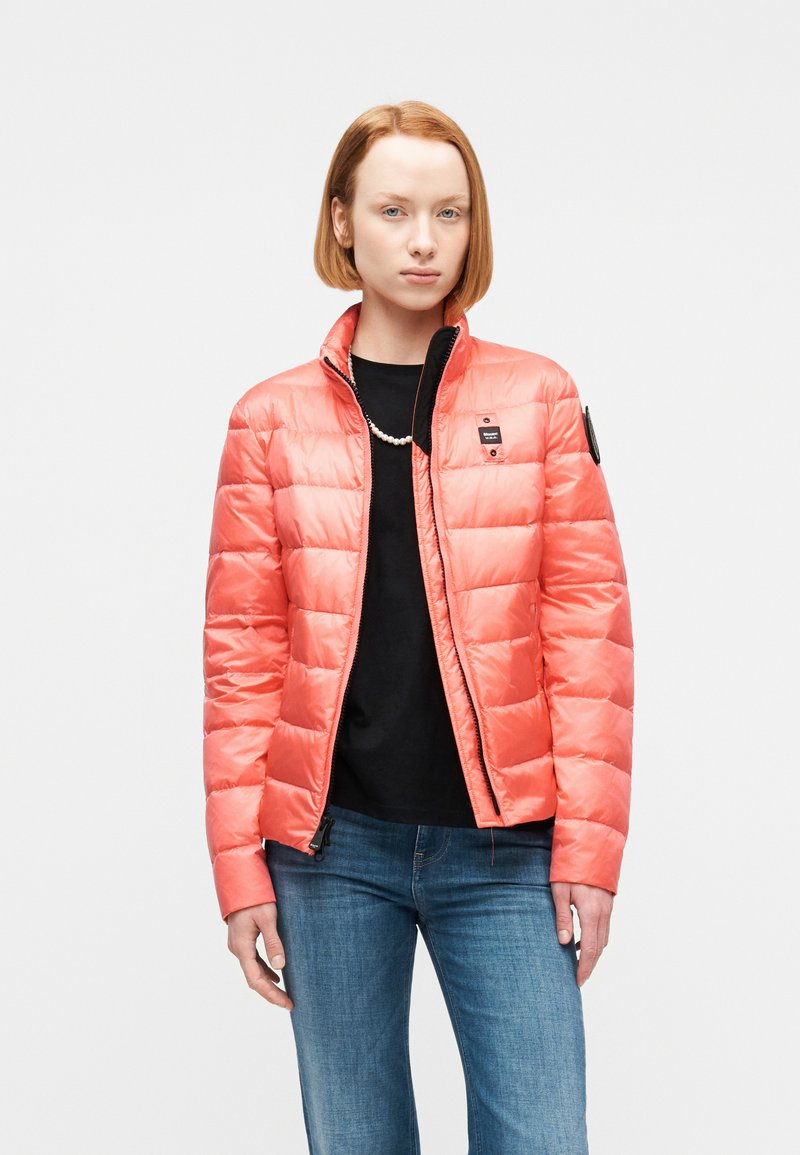 Femme aux cheveux rouges courts portant une veste matelassée corail par-dessus un chemisier noir et un jean, debout devant un fond blanc uni.